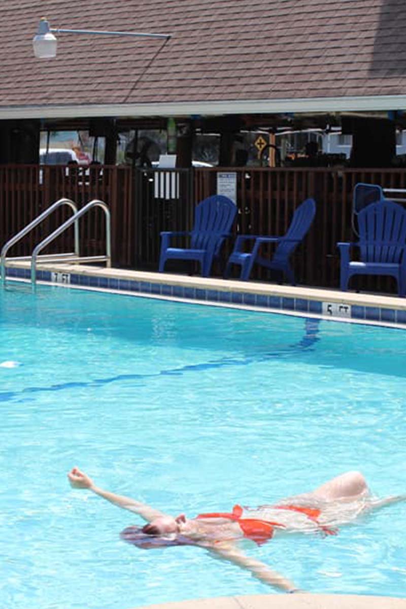 A person swimming in a pool with blue chairs and a brown roof in the background.